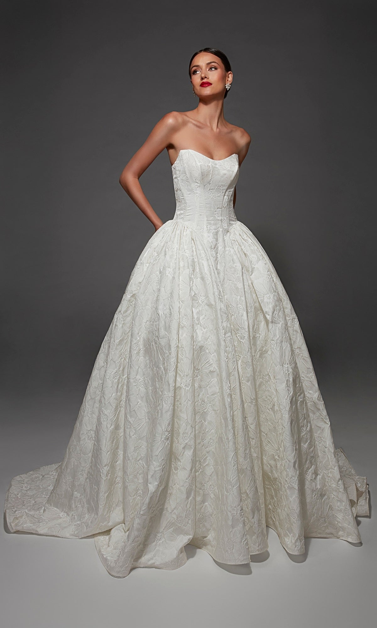 Woman wearing the Long Wedding Dress: Alyce 7174, a strapless jacquard ballgown with a fitted corset bodice and a voluminous, textured skirt, standing against a plain dark background.